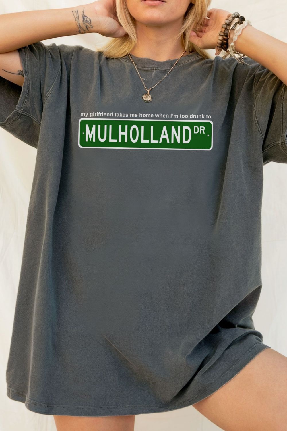 gray t-shirt with Mulholland Drive street sign design on a white background, with blink 182 lyrics my girlfriend takes me home when I'm too drunk to drive, worn by a person