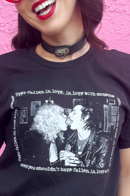 Person wearing a black t-shirt with a 70s punk graphic design and text, against a pink background.